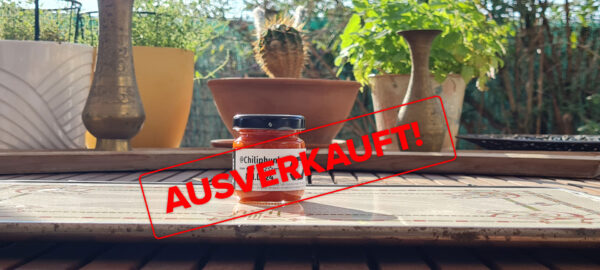 Label mit dem Text "Ausverkauft" liegt über dem Bild der Chilipaste MID24. Das Bild: 1 Glas Chilipaste Chiliphunk MID24 mit 3 Sternen am Etikett, auf einem Holztisch; Dahinter ist unscharf der Gartenzaun und Kräutertöpfe am Tisch. In der Mitte ein Kaktus, zwei kleine Messingvasen links und rechts.