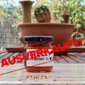 Label mit dem Text "Ausverkauft" liegt über dem Bild der Chilipaste Basis Brennt ’24. Das Bild: 1 Glas Chilipaste Chiliphunk Basis Brennt ’24 mit 3 Sternen am Etikett, auf einem Holztisch; Dahinter ist unscharf der Gartenzaun und Kräutertöpfe am Tisch. In der Mitte ein Kaktus, zwei kleine Messingvasen links und rechts.