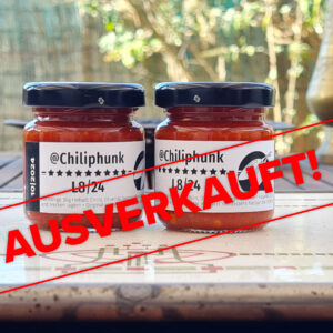 Label mit dem Text "Ausverkauft" liegt über dem Bild. Das Bild: 2 Gläser Chiliphunk L8/24 Chilipaste auf einem Holztisch; Dahinter ist unscharf der Gartenzaun und Messingvasen am Tisch.