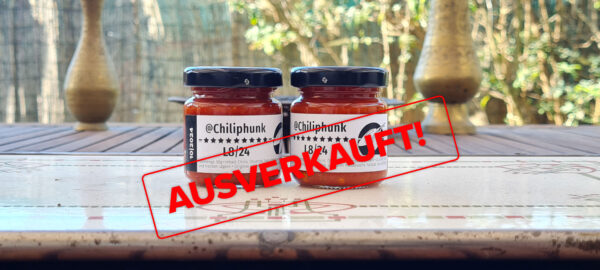 Label mit dem Text "Ausverkauft" liegt über dem Bild. Das Bild: 2 Gläser Chiliphunk L8/24 Chilipaste auf einem Holztisch; Dahinter ist unscharf der Gartenzaun und Messingvasen am Tisch.